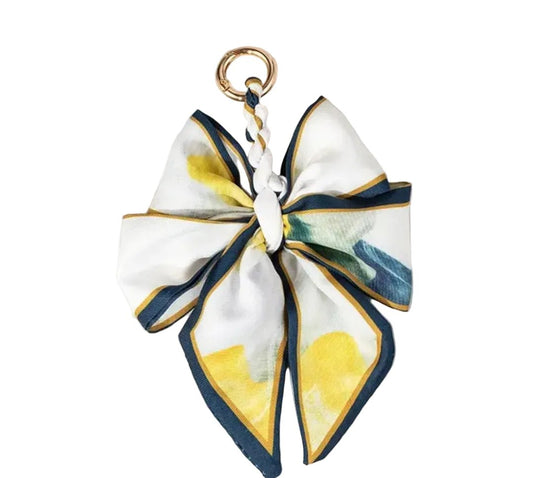 A silk scarf keychain charm shaped like a bow, featuring a white base with yellow watercolor-style floral accents, navy blue borders, and a gold metal key ring at the top.