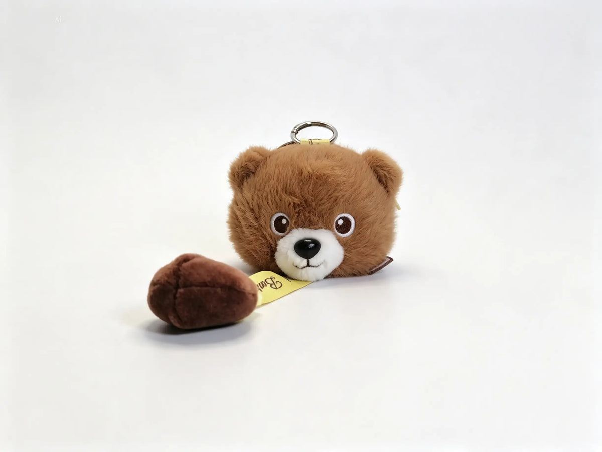 A small plush bear keychain or coin purse in medium brown with a white muzzle, large round eyes, and a metal key ring at the top. A small yellow tag is attached near the mouth, and a separate dark brown plush piece lies beside it on a clean white background.