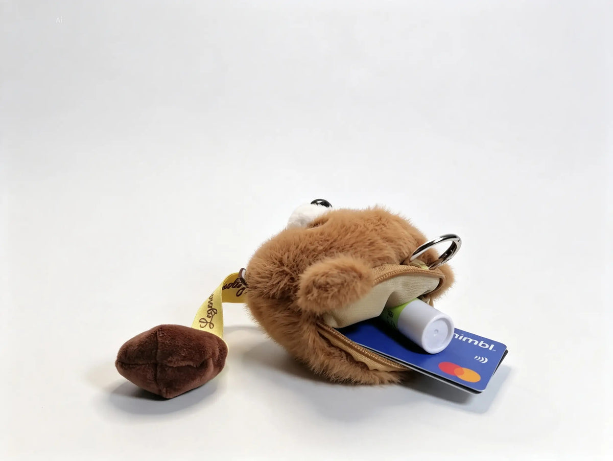 A small plush keychain or pouch in tan and brown faux fur, shown with a small brown poop-shaped accessory and a blue credit card tucked into its open pocket. 