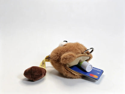 A small plush keychain or pouch in tan and brown faux fur, shown with a small brown poop-shaped accessory and a blue credit card tucked into its open pocket. 